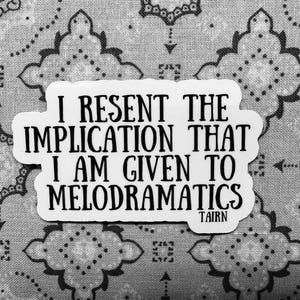 Puede incluir: Una pegatina blanca con texto negro: "I RESENT THE IMPLICATION THAT I AM GIVEN TO MELODRAMATICS" y el nombre "TAIRN" debajo. Sobre un fondo gris con estampado.
