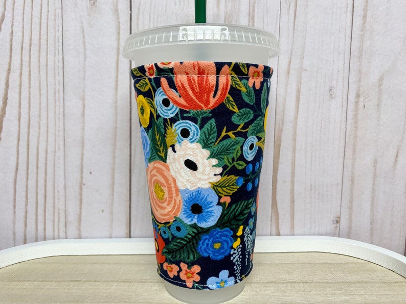 May include: A reusable cup cosy with a floral pattern in shades of blue, pink, yellow, and green on a dark blue background. The cosy is made of fabric and is designed to fit a standard-sized cup.