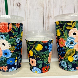 May include: Three reusable cup sleeves with a floral pattern on a dark blue background. The sleeves are made of fabric and have a sewn seam around the top edge. The sleeves are designed to fit around a standard-sized cup.