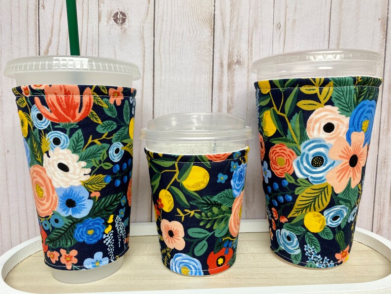 May include: Three reusable cup sleeves with a floral pattern on a dark blue background. The sleeves are made of fabric and have a sewn seam around the top edge. The sleeves are designed to fit around a standard-sized cup.
