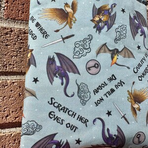May include: A blue fabric pouch with a repeating pattern of dragons, griffins, swords, keys, clouds, and stars. The pouch has the text "Be Where Needed", "Scratch Her Eyes Out", "Die Today You Will Not", and "Civility is Overrated".