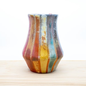 May include: A ceramic vase with a colourful striped design. The vase features vertical stripes in shades of red, orange, yellow, and blue, with metallic gold accents. The vase is approximately 20 cm tall and sits on a wooden surface.