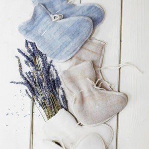 May include: Three pairs of fabric lavender sachets in blue, beige, and white. The sachets are tied with white string and are arranged on a white wooden surface with a bundle of dried lavender.