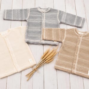 May include: Three long-sleeved button-down cardigans in cream, grey, and tan. Each cardigan features a round neckline, a front button closure, and a small rectangular label at the bottom hem. The grey and tan cardigans have a subtle horizontal striped pattern.