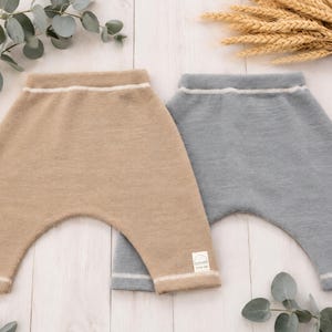 May include: Two pairs of knit trousers, one in tan and one in slate blue, featuring a high-waisted design with a thin white horizontal stripe at the waistband and cuffs. Each pair includes a small white rectangular label with a rainbow icon and text.