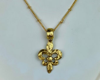 May include: A gold necklace with a fleur-de-lis pendant. The chain is made of small gold beads.