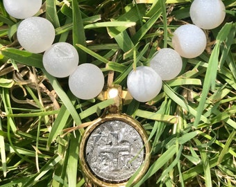 May include: White and gold beaded bracelet with a silver coin charm. The beads are made of white agate and coral. The coin charm has a cross design.