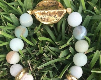 May include: A bracelet made of light green and white stones with a gold charm in the center. The bracelet is on a bed of green grass.