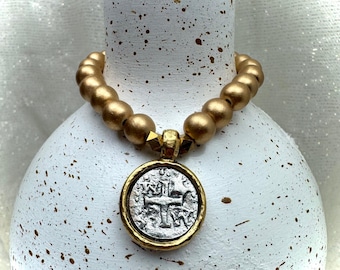 May include: A gold beaded necklace with a silver coin pendant featuring a cross design. The coin is set in a gold frame. The necklace is displayed on a white vase with gold speckles.