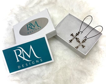 May include: A pair of silver cross earrings with black wire hooks, displayed in a white box with the logo "RM Designs" on the lid and a teal box with the same logo on the bottom.