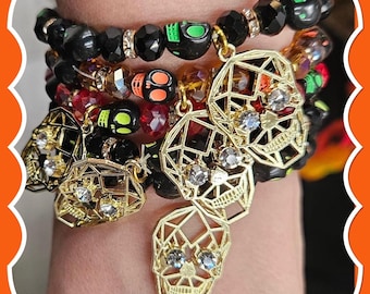 May include: A multi-strand stretch bracelet featuring black faceted beads, translucent red beads, and small skull-shaped beads in neon green and orange. Several gold-tone geometric skull charms are attached, each accented with clear rhinestones.