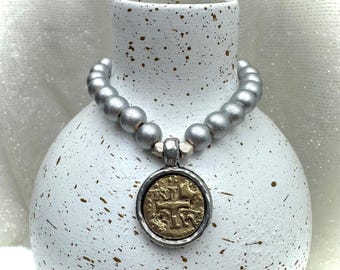 May include: A silver beaded necklace with a circular pendant featuring a gold coin design. The necklace is draped around a white vase with brown speckles, creating a vintage aesthetic.