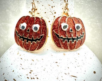 May include: A pair of Halloween-themed earrings. Each earring is shaped like a red glitter pumpkin with a black carved face and gold-colored trim. The eyes are silver-colored and the earrings hang from gold-colored hooks.
