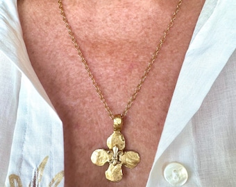 May include: A gold-toned necklace featuring a textured, four-petaled cross pendant with a central fleur-de-lis emblem. The pendant hangs from a delicate gold-toned chain, resting against a white collared shirt with a visible button.