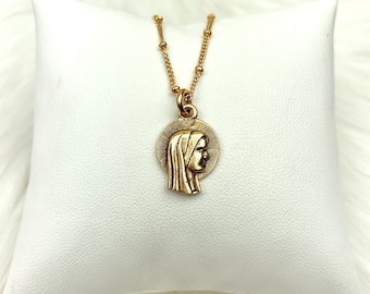 May include: A gold chain necklace with a pendant featuring a profile of the Virgin Mary. The pendant is circular and has a halo around the Virgin Mary's head.