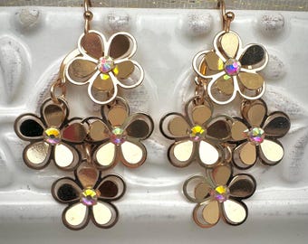 May include: A pair of gold-toned flower earrings. Each earring features three cascading flower charms with iridescent centers. The earrings have a hook closure and are designed with a layered, openwork style, creating a delicate and feminine aesthetic.