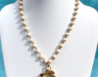 May include: A white beaded necklace with a gold flower pendant. The necklace is on a white mannequin.
