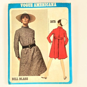 May include: A vintage Vogue Americana sewing pattern featuring a design by Bill Blass. The pattern is for a dress and coat, and the cover shows a model wearing a grey dress with a black belt and a white hat, and another model wearing a red coat.