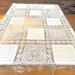 May include: A large, rectangular white lace tablecloth with a patchwork design. The tablecloth features square and rectangular panels with various embroidered patterns and a scalloped edge. The lace is detailed and intricate, creating a vintage aesthetic.