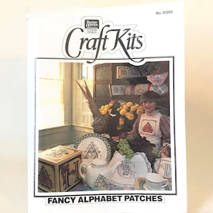 May include: A craft kit featuring a set of alphabet patches. The image shows a girl holding a patch with a letter A, surrounded by other patches, a teapot, and a pheasant. The kit includes instructions for making alphabet patches.