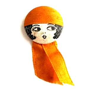 May include: Orange fabric button with a hand-painted design of a woman with black hair wearing a hat. The button has a long orange fabric tail.