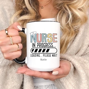 May include: White ceramic mug with a black handle and a black rim. The mug has a colorful graphic that says "Nurse in Progress... Loading... Please Wait" with a progress bar. The name "Katie" is printed below the progress bar.