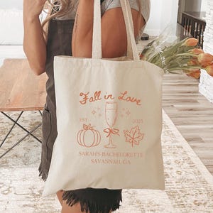 May include: Cream-colored tote bag with the text "Fall in Love" and a pumpkin, champagne glass, and leaf graphic in orange. The text "SARAH'S BACHELORETTE SAVANNAH, GA" is also printed on the bag. The bag has long, cream-colored handles.