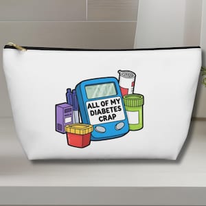 May include: White zippered cosmetic bag with a black trim, featuring a blue blood glucose meter, medicine bottles, and the text "ALL OF MY DIABETES CRAP". The bag is on a white surface.