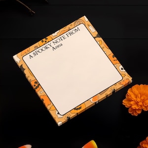 May include: A Halloween-themed notepad with the text "A Spooky Note From Anna" on a black surface. The notepad is surrounded by orange pumpkins, candy corn, and orange flowers. A bare branch is in the background.