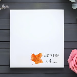 May include: A white notepad with the text "A Note From Anna" and a watercolor orange leaf illustration. The notepad is surrounded by a pink rose, eucalyptus leaves, and lavender sprigs on a dark gray wooden surface.