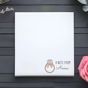 May include: A white square notepad with a ring graphic and the text "A note from Anna". The ring graphic has a diamond, a bow, and a gold band. The notepad is surrounded by a pink rose, lavender, and eucalyptus leaves.