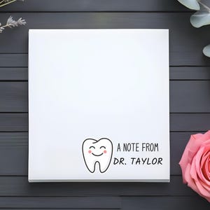 May include: A white card with a cartoon tooth with a smiling face and the text "A note from Dr. Taylor". The card is on a dark gray wooden surface with a sprig of lavender and a sprig of eucalyptus on either side of the card. A pink rose is on the right side of the card.