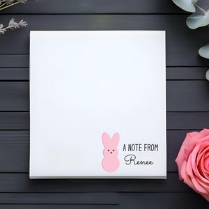 May include: A white notepad with a pink bunny illustration and the text "A note from Renee". The notepad is on a dark wood surface with a pink rose and green foliage.