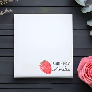 May include: A white notepad with a red strawberry illustration and the text "A note from Amelia". The notepad is on a dark wood surface with lavender and eucalyptus sprigs.
