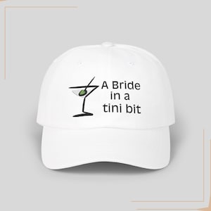 May include: White baseball cap with black embroidery of a martini glass with an olive and the text "A Bride in a Tini Bit".