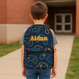 May include: A navy blue backpack with a pattern of colorful video game controllers. The name "Aidan" is printed in yellow on the front. The backpack has a black zipper and a mesh side pocket.