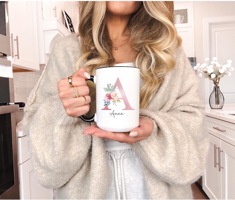 A white ceramic mug with a black rim and handle. The mug has a pink floral design with the letter 'A'. Mug can be personalized with name under initial. Perfect personalize gift.