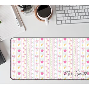 K&ouml;nnte beinhalten: Rechteckige Schreibtischunterlage mit wei&szlig;em Hintergrund und einem sich wiederholenden Muster aus rosa &Auml;pfeln, gelben Bleistiften, Schleifen und Sternen. Die Matte hat vertikale Streifen in Rosa, Gelb und Blau. Der Name "Mrs. Smith" ist kursiv geschrieben.
