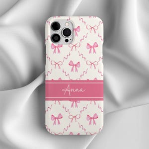 Puede incluir: Carcasa de teléfono blanca con un patrón de lazos rosa y blanco y el nombre "Anna" impreso en blanco sobre una banda rosa.