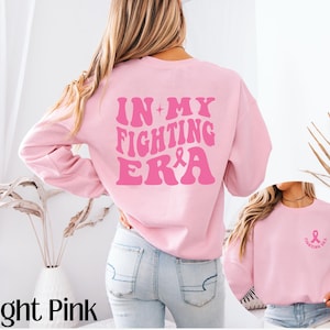 May include: Light pink sweatshirt with the text "IN MY FIGHTING ERA" in pink with a pink ribbon graphic. The sweatshirt has a crew neck and long sleeves.