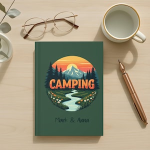 May include: A green journal with a circular illustration of a mountain landscape with the word "CAMPING" in orange and white. The journal also has the names "Mark & Anna" printed on it. A pair of glasses, a pen, and a mug are also on the wooden surface.
