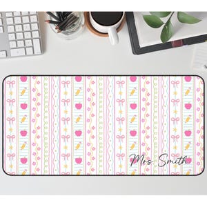K&ouml;nnte beinhalten: Rechteckige Schreibtischunterlage mit einem sich wiederholenden Muster aus rosa &Auml;pfeln, gelben Bleistiften und rosa Schleifen auf wei&szlig;em Hintergrund. Die Matte tr&auml;gt den Text "Mrs. Smith" in Schreibschrift.
