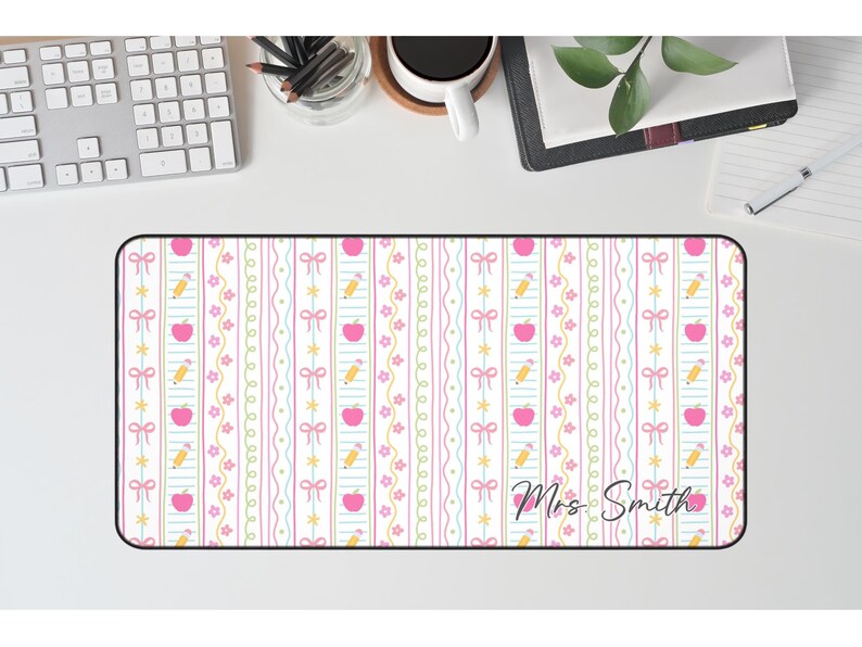 K&ouml;nnte beinhalten: Rechteckige Schreibtischunterlage mit einem sich wiederholenden Muster aus rosa &Auml;pfeln, gelben Bleistiften und rosa Schleifen auf wei&szlig;em Hintergrund. Die Matte tr&auml;gt den Text "Mrs. Smith" in Schreibschrift.