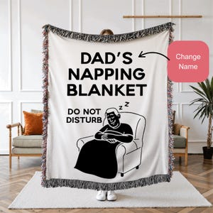 May include: A white woven blanket with black fringe and the text "DAD'S NAPPING BLANKET" and "DO NOT DISTURB". A cartoon illustration of a person napping in a chair is also on the blanket. The blanket is being held up by a person.