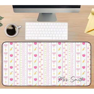 K&ouml;nnte beinhalten: Rechteckige Schreibtischunterlage mit wei&szlig;em Hintergrund und einem sich wiederholenden Muster aus rosa &Auml;pfeln, Bleistiften, Schleifen und Sternen. Die Matte hat vertikale Streifen in Rosa und Gr&uuml;n. Der Name "Mrs Smith" ist kursiv geschrieben.