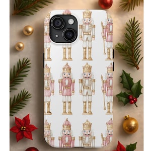 May include: A white phone case featuring a repeating pattern of watercolor-style nutcracker soldiers in pink, gold, and beige. The case has a black camera module and is decorated with Christmas ornaments and greenery.
