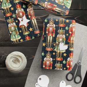 May include: Gift-wrapped boxes with a festive nutcracker design. The wrapping paper features illustrations of nutcrackers, stars, and ornaments on a dark blue background. A spool of white twine, scissors, and gift tags are also visible.