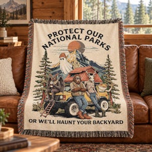 May include: A woven throw blanket featuring a graphic of a yellow off-road vehicle, a ghost, a moth-like creature, a sasquatch, and a deer-headed figure. The text reads PROTECT OUR NATIONAL PARKS OR WE'LL HAUNT YOUR BACKYARD.