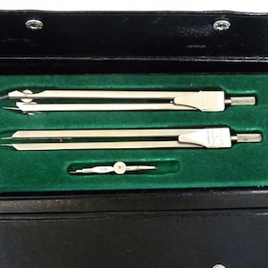 May include: A set of three silver compasses in a black leather case with a green velvet lining. The compasses are in a closed position.