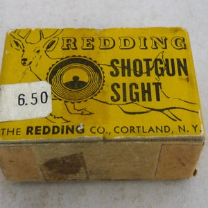 May include: A yellow cardboard box with black text that reads "REDDING SHOTGUN SIGHT" and "THE REDDING CO., CORTLAND, N.Y.". The box has a price tag that reads "6.50". The box features a black and white illustration of a deer.